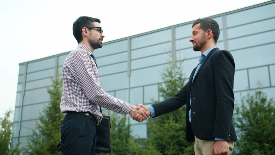 Two businessmen in casual attire shaking hands outside a modern office building.